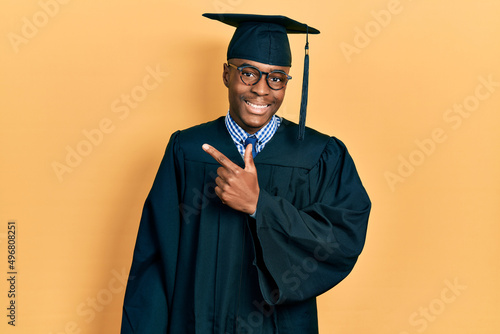 Young african american man wearing graduation cap and ceremony robe cheerful with a smile on face pointing with hand and finger up to the side with happy and natural expression photo