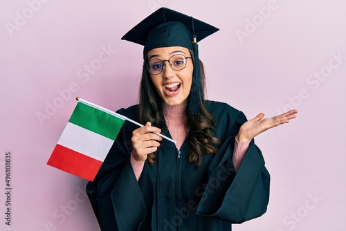 Young hispanic woman wearing graduation uniform holding italy flag celebrating achievement with happy smile and winner expression with raised hand