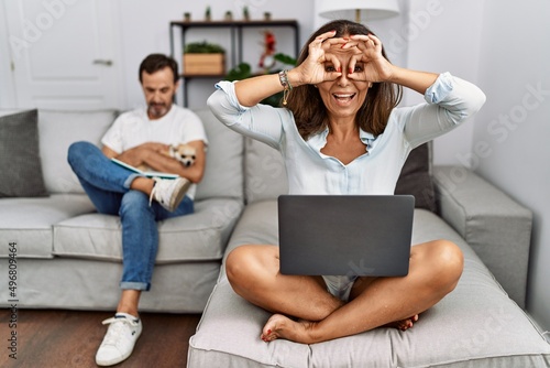 Hispanic middle age couple at home, woman using laptop doing ok gesture like binoculars sticking tongue out, eyes looking through fingers. crazy expression.