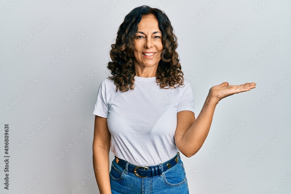 Middle age hispanic woman wearing casual white t shirt smiling cheerful presenting and pointing with palm of hand looking at the camera. Middle age hispanic woman wearing casual white t shirt smiling cheerful presenting and pointing with palm of hand looking at the camera.