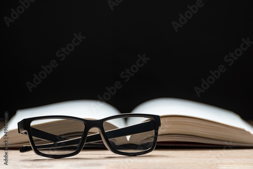 Glasses for vision and an open book lie on a table on a black background. Reading Glasses. Classic black frame glasses.