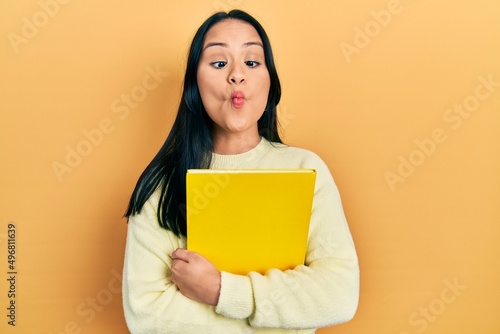 Beautiful hispanic woman with nose piercing holding book making fish face with mouth and squinting eyes, crazy and comical.