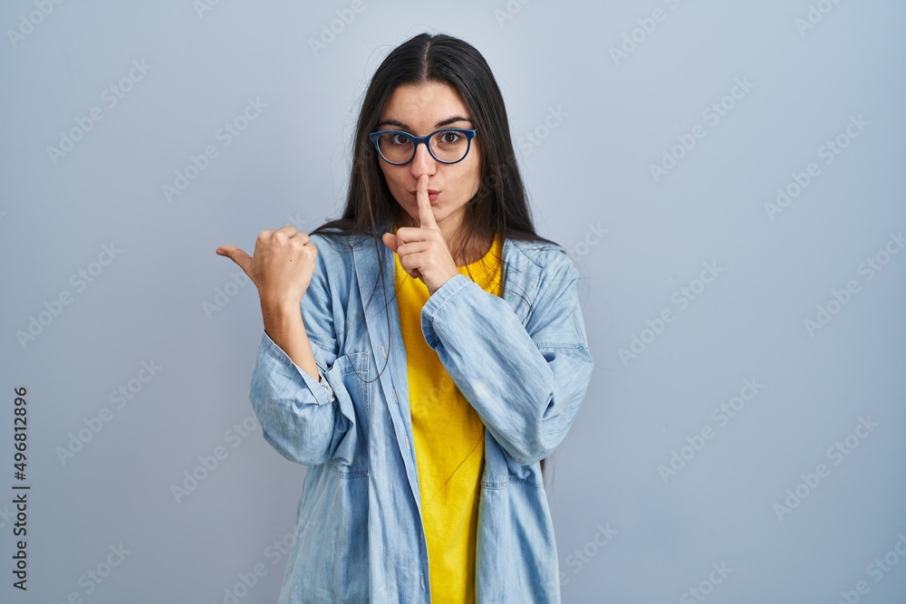 Young hispanic woman standing over blue background asking to be quiet with finger on lips pointing with hand to the side. silence and secret concept. Young hispanic woman standing over blue background asking to be quiet with finger on lips pointing with hand to the side. silence and secret concept.