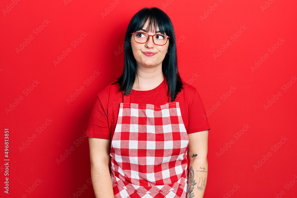 Young hispanic woman wearing cook apron and glasses smiling looking to the side and staring away thinking.
