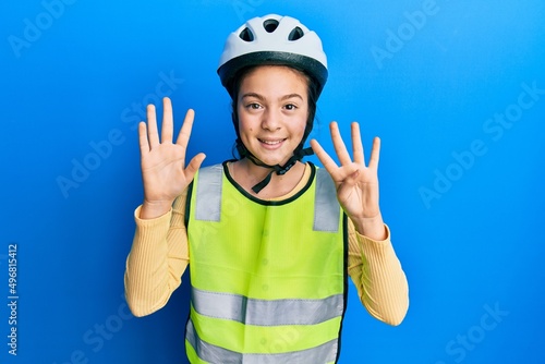 Beautiful brunette little girl wearing bike helmet and reflective vest showing and pointing up with fingers number nine while smiling confident and happy.