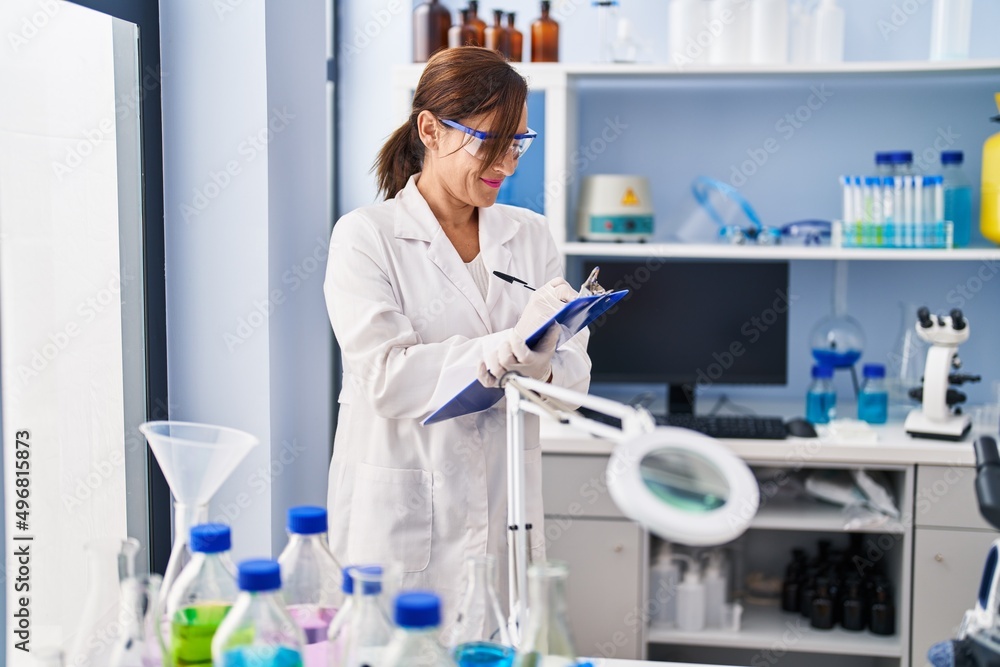 Middle age woman wearing scientist uniform writing on clipboard at laboratory Middle age woman wearing scientist uniform writing on clipboard at laboratory