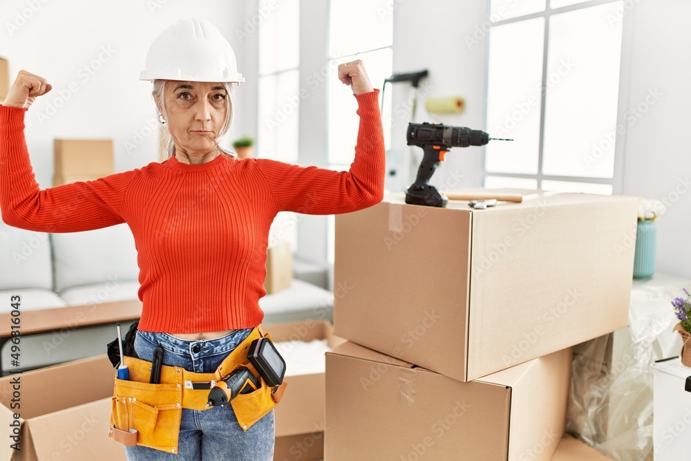Middle age grey-haired woman wearing hardhat standing at new home showing arms muscles smiling proud. fitness concept. Middle age grey-haired woman wearing hardhat standing at new home showing arms muscles smiling proud. fitness concept.