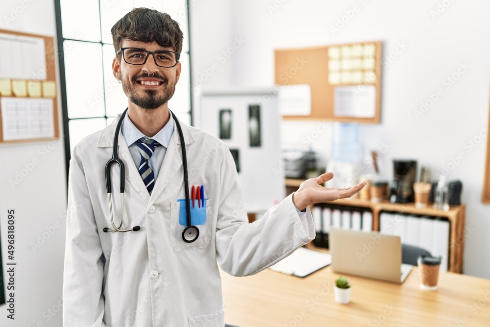 Hispanic man with beard wearing doctor uniform and stethoscope at the office smiling cheerful presenting and pointing with palm of hand looking at the camera. Hispanic man with beard wearing doctor uniform and stethoscope at the office smiling cheerful presenting and pointing with palm of hand looking at the camera.