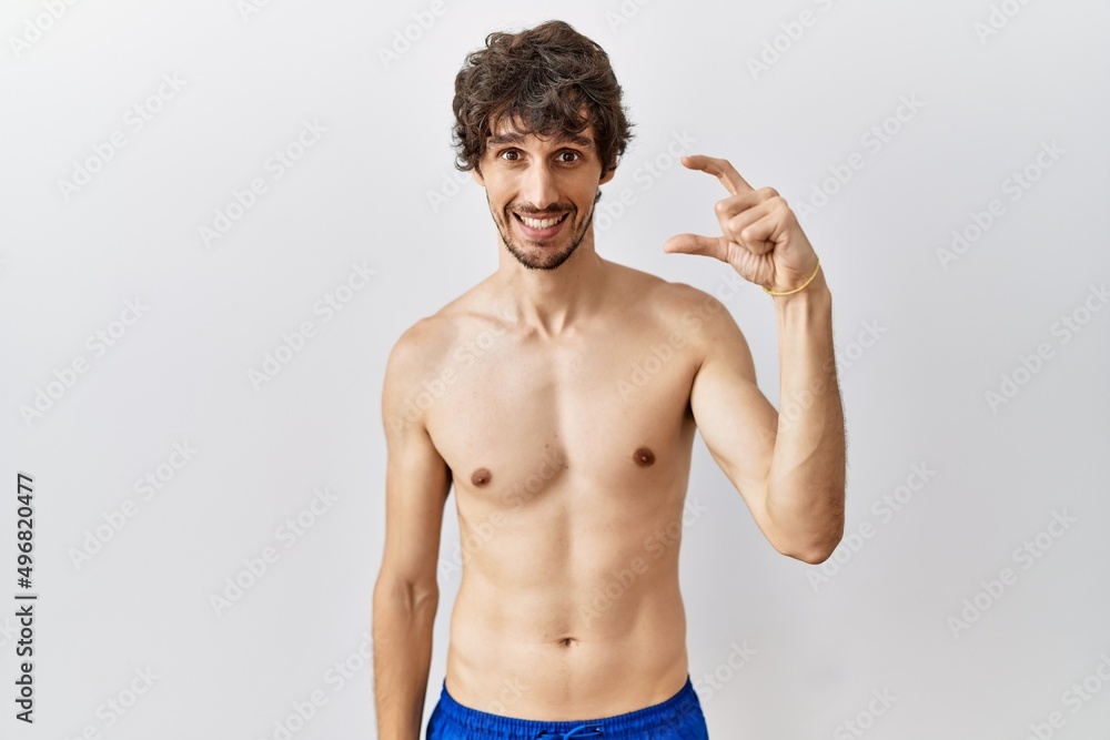 Fototapeta premium Young hispanic man standing shirtless over isolated, background smiling and confident gesturing with hand doing small size sign with fingers looking and the camera. measure concept.