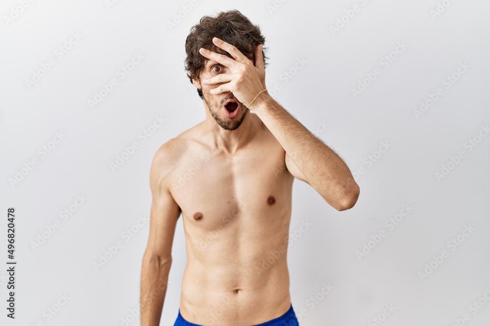Fototapeta premium Young hispanic man standing shirtless over isolated, background peeking in shock covering face and eyes with hand, looking through fingers with embarrassed expression.