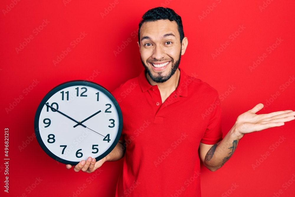 Hispanic man with beard holding big clock celebrating achievement with happy smile and winner expression with raised hand Hispanic man with beard holding big clock celebrating achievement with happy smile and winner expression with raised hand