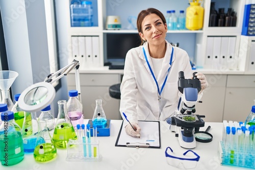 Young hispanic woman wearing scientist uniform using microscope and writing on clipboard at laboratory