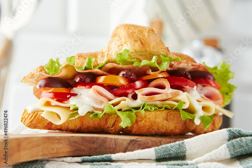 Croissant sandwich with cheese, ham, lettuce, tomatoes, olives and delicious sauce on wooden board.