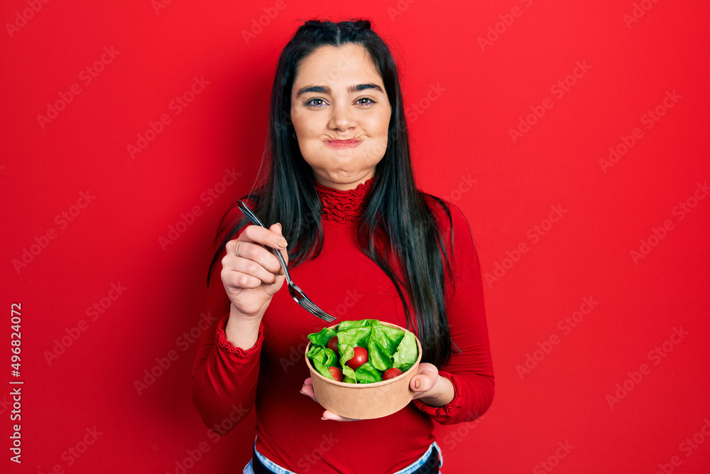 Young hispanic girl eating salad puffing cheeks with funny face. mouth inflated with air, catching air. Young hispanic girl eating salad puffing cheeks with funny face. mouth inflated with air, catching air.