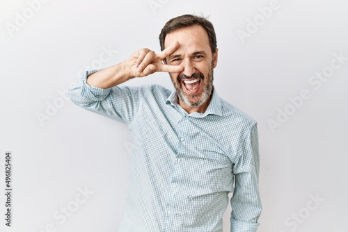 Middle age hispanic man with beard standing over isolated background doing peace symbol with fingers over face, smiling cheerful showing victory © Krakenimages.com