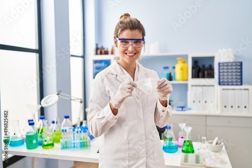 Young blonde woman wearing scientist uniform holding medical mask at laboratory