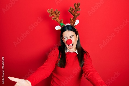 Young hispanic woman wearing deer christmas hat and red nose smiling cheerful offering hands giving assistance and acceptance.