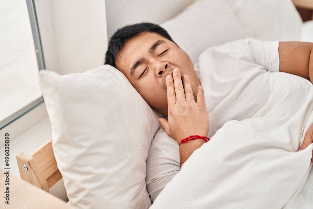 Young chinese man lying on bed yawning at bedroom
