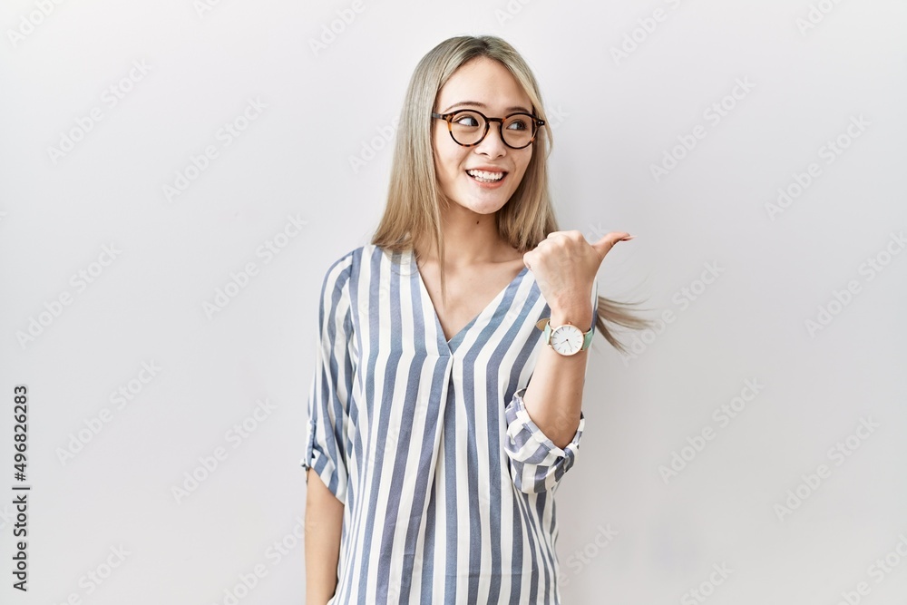Asian young woman wearing casual clothes and glasses smiling with happy face looking and pointing to the side with thumb up. Asian young woman wearing casual clothes and glasses smiling with happy face looking and pointing to the side with thumb up.