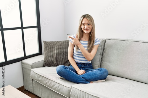 Asian young woman sitting on the sofa at home cheerful with a smile of face pointing with hand and finger up to the side with happy and natural expression on face