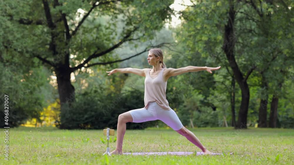 Young woman doing yoga exercise in front of device in green park on summer spbi. 4k European female does asana or gymnastics and stretches her body, looks with smile and stands on mat in front of Young woman doing yoga exercise in front of device in green park on summer spbi. 4k European female does asana or gymnastics and stretches her body, looks with smile and stands on mat in front of