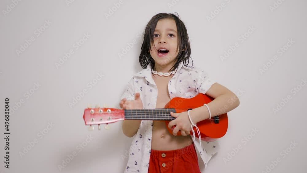 Musician Hawaiian child boy with long hair, shirt with a red small guitar playing against a white wall Musician Hawaiian child boy with long hair, shirt with a red small guitar playing against a white wall
