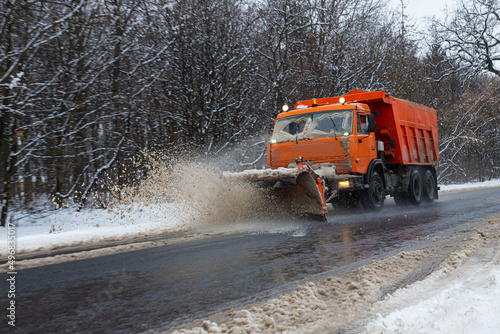 A large car with a plow clears the road from snow. Orange cargo special equipment is struggling with the elements in winter. Removing the effects of the snowstorm. Difficulties in traffic.Frozen water