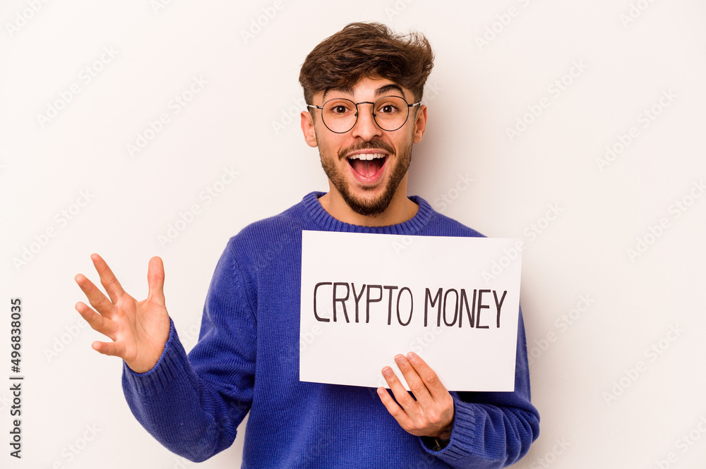 Young hispanic man holding a crypto money placard isolated on white background receiving a pleasant surprise, excited and raising hands. Young hispanic man holding a crypto money placard isolated on white background receiving a pleasant surprise, excited and raising hands.