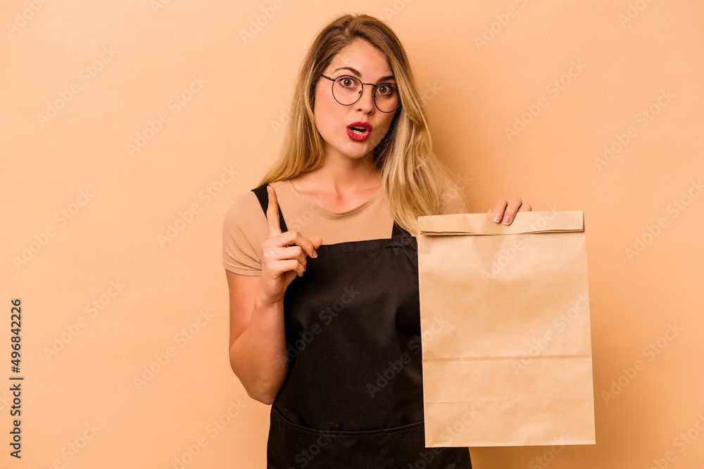 Restaurant waiter caucasian woman holding a take away bag isolated on beige background having some great idea, concept of creativity. Restaurant waiter caucasian woman holding a take away bag isolated on beige background having some great idea, concept of creativity.
