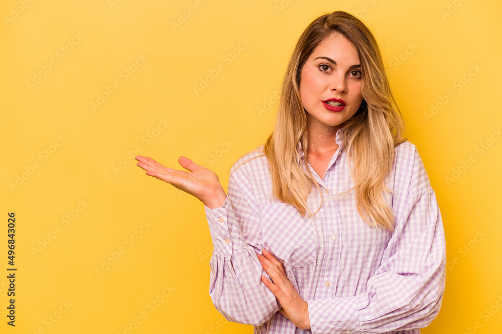 Young caucasian woman isolated on yellow background impressed holding copy space on palm. Young caucasian woman isolated on yellow background impressed holding copy space on palm.