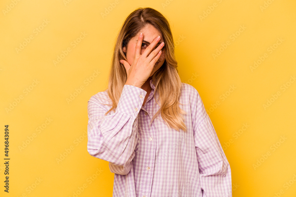 Young caucasian woman isolated on yellow background blink at the camera through fingers, embarrassed covering face. Young caucasian woman isolated on yellow background blink at the camera through fingers, embarrassed covering face.