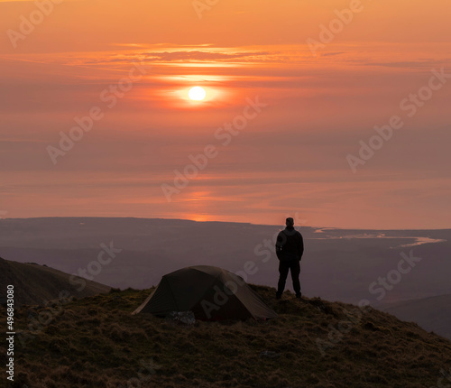 Silhouette of an adventurer and wild camping tent high in the mountains at sunset