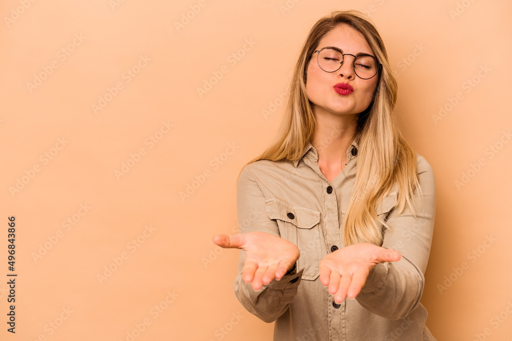 Young caucasian woman isolated on beige background folding lips and holding palms to send air kiss. Young caucasian woman isolated on beige background folding lips and holding palms to send air kiss.