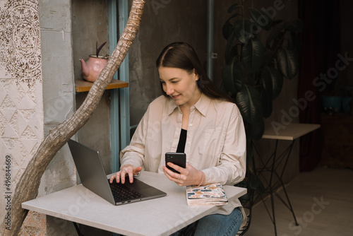 A young European freelancer girl with long dark hair in a shirt, jeans and sneakers remotely works in a cafe, businesswoman are working on laptop and smiling