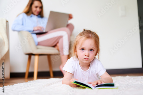 Cute little girl reading a book on the floor at home while mother using laptop