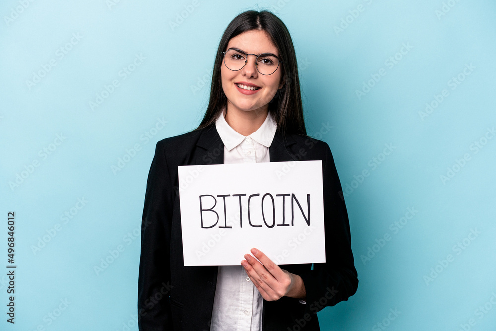 Young caucasian business woman holding a bitcoin placard isolated on blue background happy, smiling and cheerful. Young caucasian business woman holding a bitcoin placard isolated on blue background happy, smiling and cheerful.