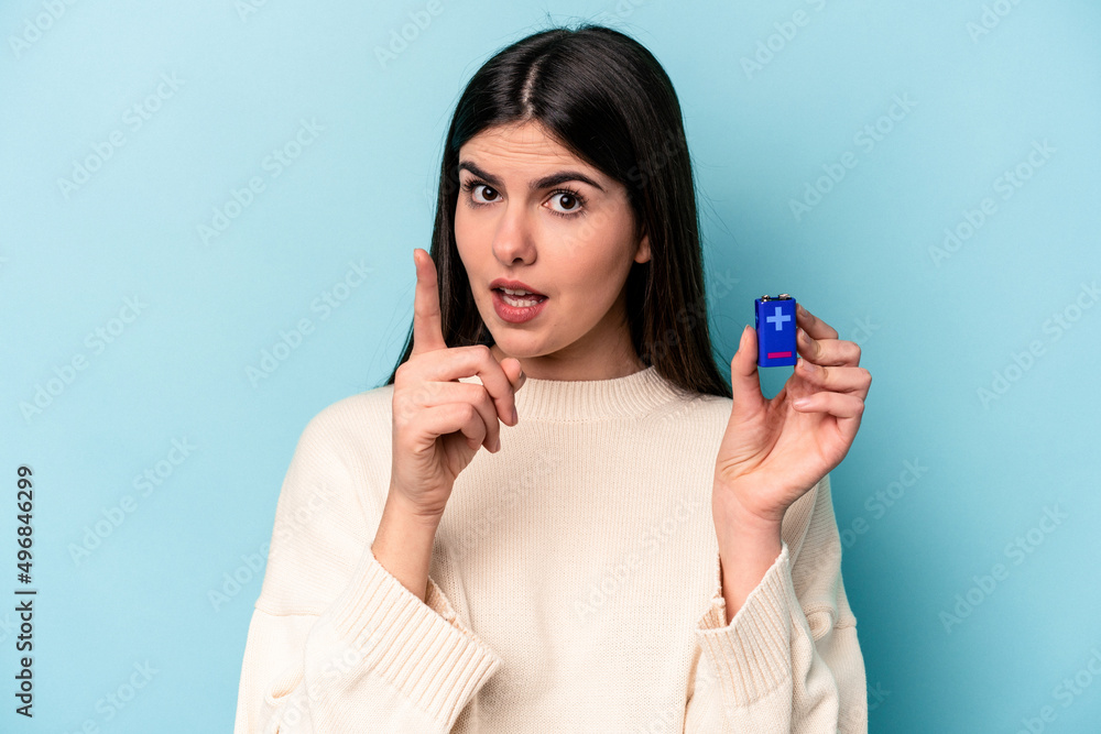 Young caucasian woman holding a batterie isolated on blue background having an idea, inspiration concept. Young caucasian woman holding a batterie isolated on blue background having an idea, inspiration concept.