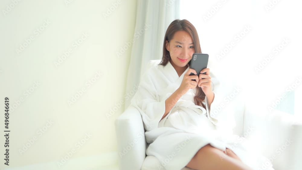 Happy asian woman in bathrobe sitting by window and checking messages on mobile phone on bright morning Happy asian woman in bathrobe sitting by window and checking messages on mobile phone on bright morning