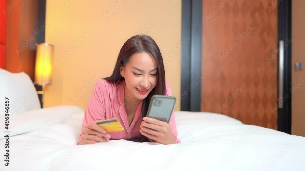 Asian woman in pink pajamas lying across bed with white comforter looks at phone and debit or credit card smiling. Online shopping, consumer debt or holiday happiness ecommerce concept. Asian woman in pink pajamas lying across bed with white comforter looks at phone and debit or credit card smiling. Online shopping, consumer debt or holiday happiness ecommerce concept.