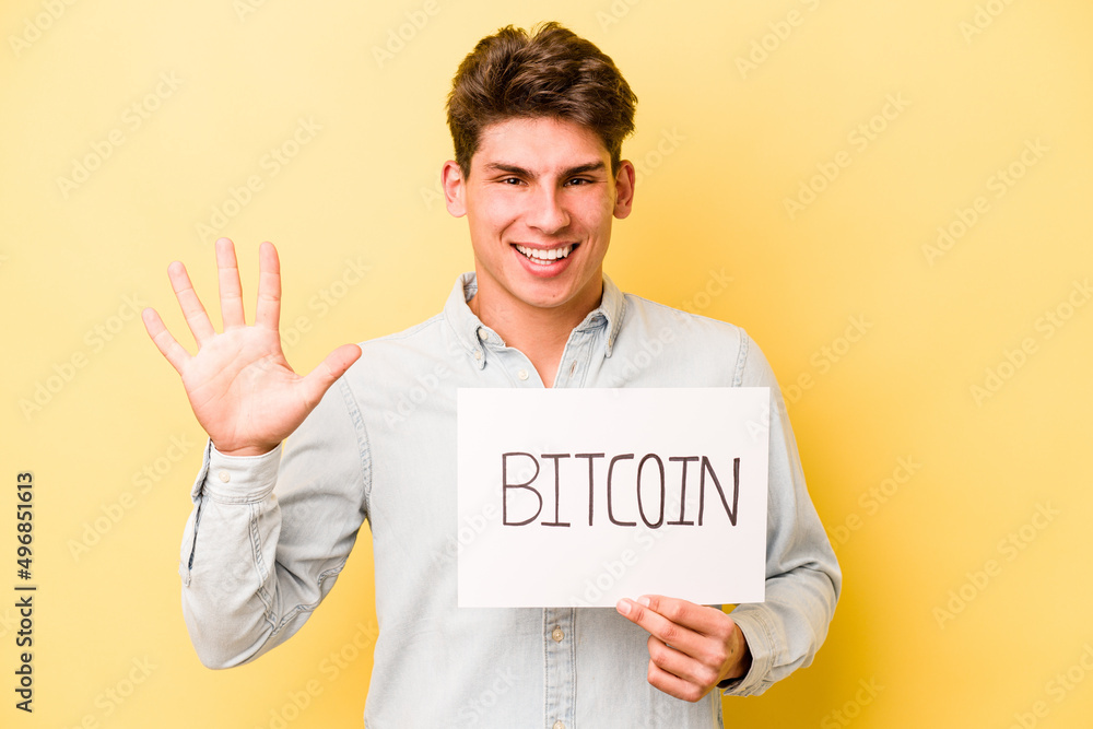 Young caucasian man holding bitcoin placard isolated on yellow background smiling cheerful showing number five with fingers. Young caucasian man holding bitcoin placard isolated on yellow background smiling cheerful showing number five with fingers.