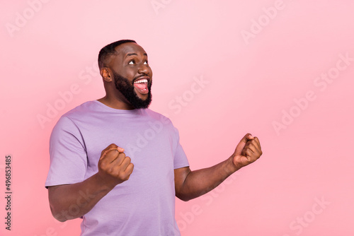 Photo of lucky excited young guy wear purple t-shirt rising fists looking up empty space isolated pink color background