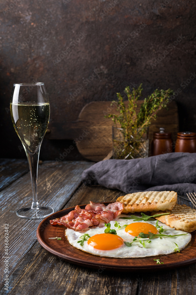 Fried eggs with bacon and herbs, 2 white bread toasts. On the table is a napkin, spices, a glass of champagne. Dark brown wooden background. Breakfast menu for cafe, restaurant, bar.