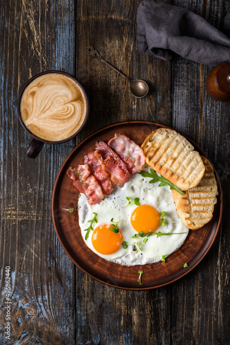 Fried eggs with bacon and herbs, 2 white bread toasts. On the table is a napkin, spices, a glass of champagne. Dark brown wooden background. Breakfast menu for cafe, restaurant, bar.