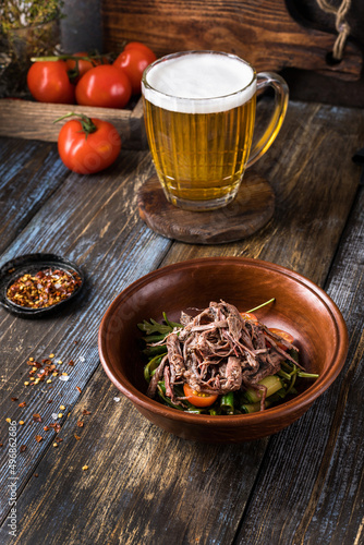 Deep plate with hot vegetables and beef. On a wooden table is a mug of light beer, spices and tomatoes. Menu for bar, cafe, restaurant. Dish from the chef. Close up