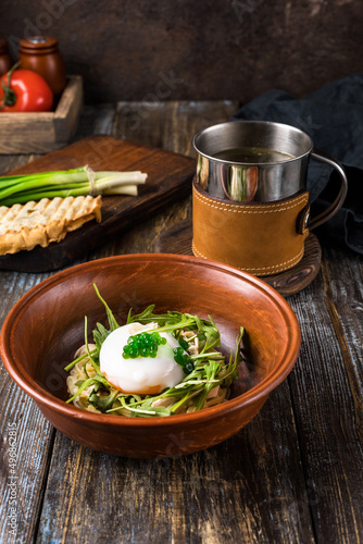 Chicken soup with arugula, chicken, poached egg in a deep bowl. On a wooden table are toasted bread, fresh onions, a mug of broth. For the restaurant menu. Close up.