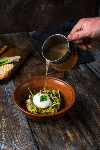 Chicken soup with arugula, chicken, poached egg in a deep bowl. On a wooden table are toasted bread, fresh onions, a mug of broth. A man's hand pours broth into a bowl. Restaurant menu. Close up.