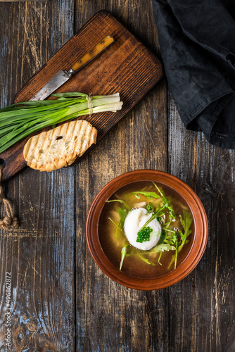 Chicken soup with arugula, chicken, poached egg in a deep bowl. On a wooden table are toasted bread, fresh onions, a mug of broth. For the restaurant menu. Top view