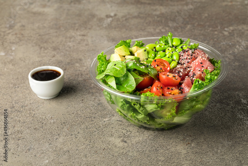 Poke bowl with tuna, salad leafs, edamame beans, avocado, cherry tomatoes and spinach on a gray textured background. Next to  soy sauce. Plastic tableware for delivery. Copy space. Close up. photo