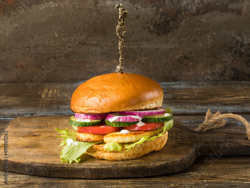 Chicken burger with tomatoes, onions, cucumbers. On the cutting board. A curly knife is stuck in a bun Brown wooden background. Close up. photo