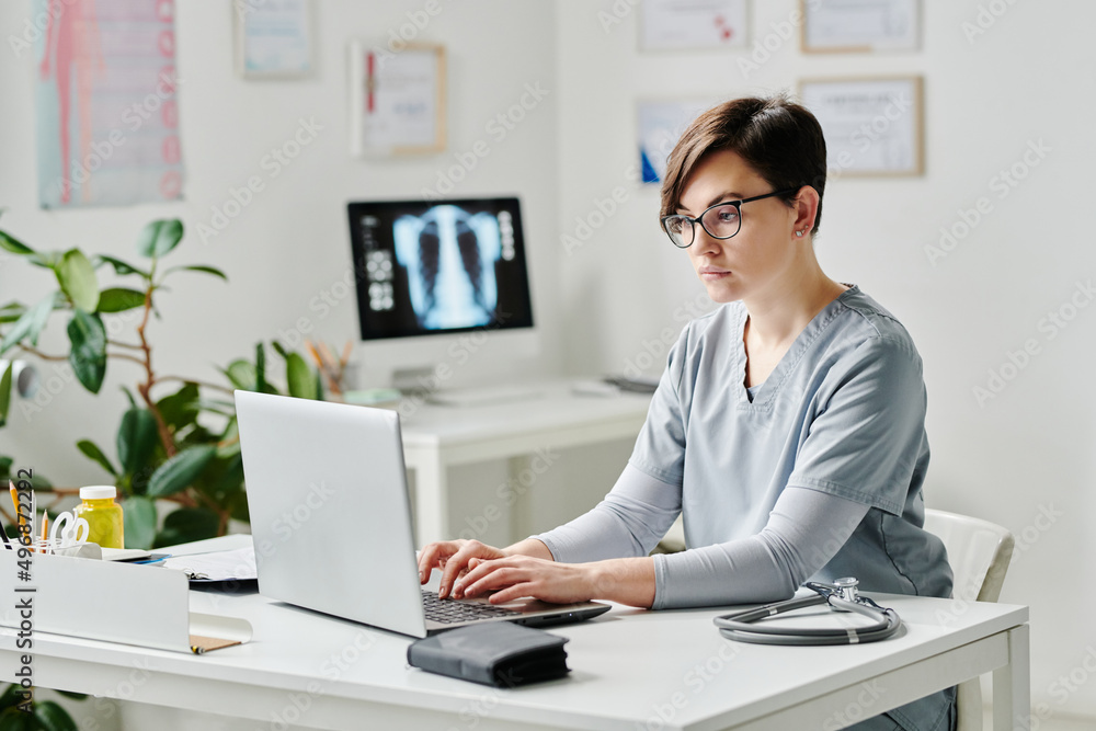 Young serious female doctor in medical scrubs typing on laptop keyboard while consulting online patient by workplace in office Young serious female doctor in medical scrubs typing on laptop keyboard while consulting online patient by workplace in office